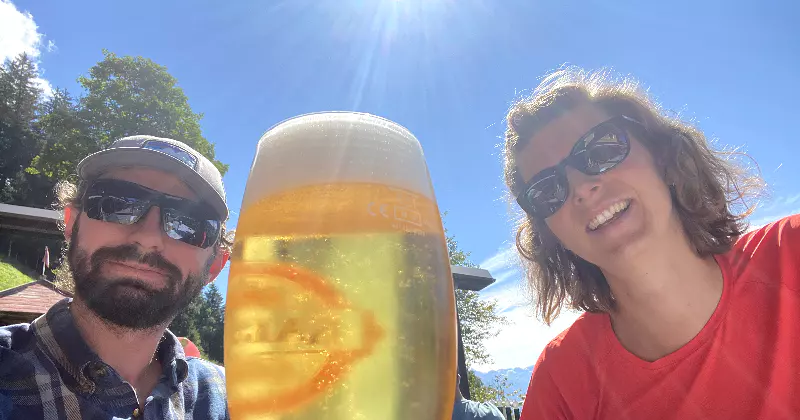 Twee mensen met een zonnebril glimlachen buiten op een zonnige dag, terwijl ze op de voorgrond zitten met een groot glas bier. Op de achtergrond zijn een blauwe lucht en groene bomen te zien.